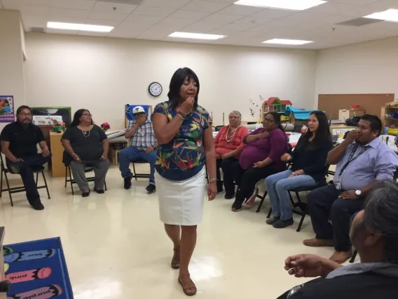 A woman with short brown hair stands in the middle of a circle of people sitting down