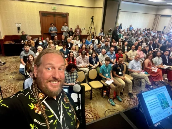 Group of people in classroom in background as blonde haired professor with beard takes a group selfie