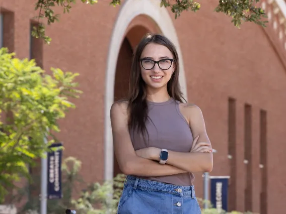 Young woman with long brown hair and glasses, stands in front of a brick building 