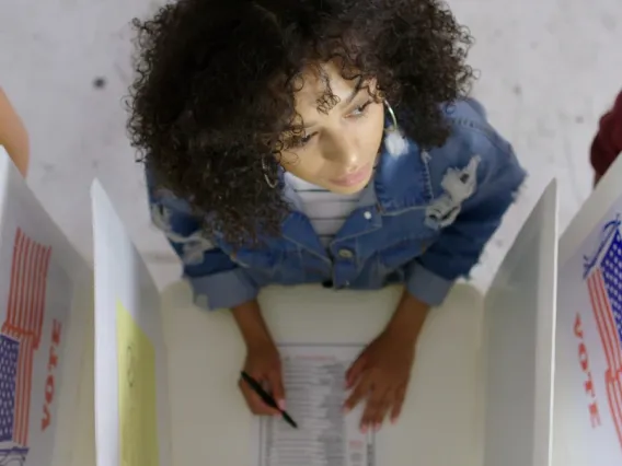 A young voter stands in a voting booth, looking up as if thinking 