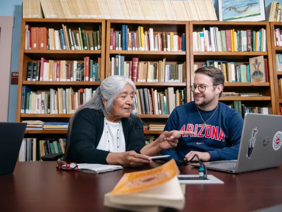 A professor and student sit together at a table in front of bookshelves