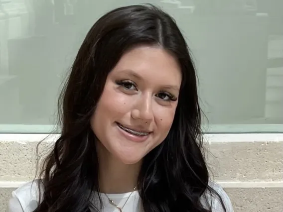 Student with long dark hair and wearing a white tshirt, smiles for the camera