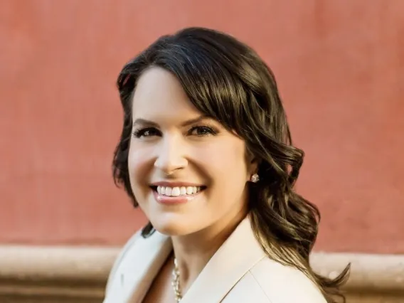 Woman with long brown hair and a white jacket stands outside smiling