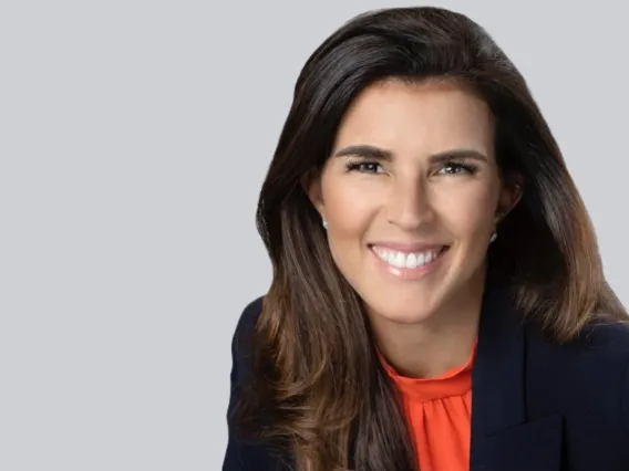 Woman with long brown hair, black jacket and orange blouse smiles in front of a gray background