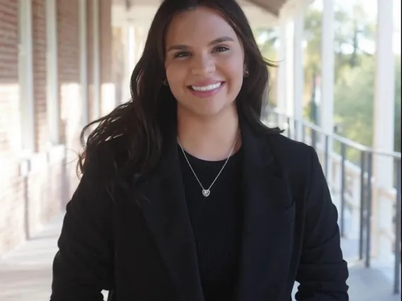 Student with long dark hair and a black outfit stands outside under a covered porch, smiling