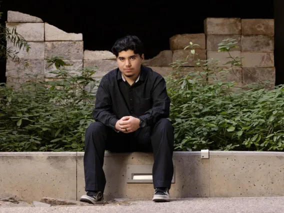 Young man with black hair and wearing all black, sits outside on a low concrete wall