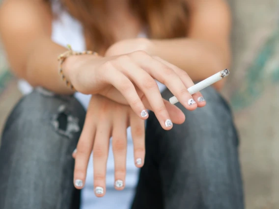 A teenager with white nail polish on their fingers, sits on a step and holds a cigarette