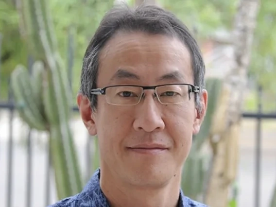 Man with dark hair, glasses and blue shirt stands outside in a wooded area