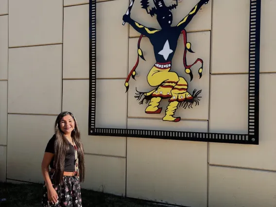 Student in Indigenous dress stands in front of an Indigenous piece of outdoor art