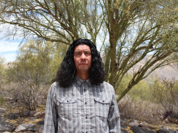 Man with long black hair and wearing a gray plaid shirt stands in front of a tree outside