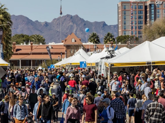 Crowd of people mill around white tents with mountains and palm trees in the background