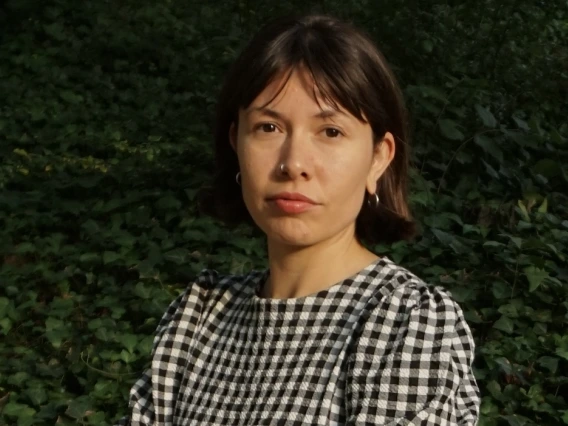 A woman with dark brown hair and wearing a black and white checked dress, stands outside with arms folded