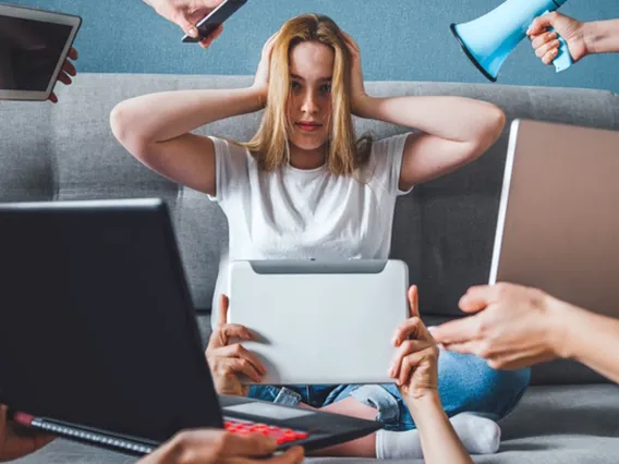 young woman sitting, hands over her ears, with several computers being placed in front of her