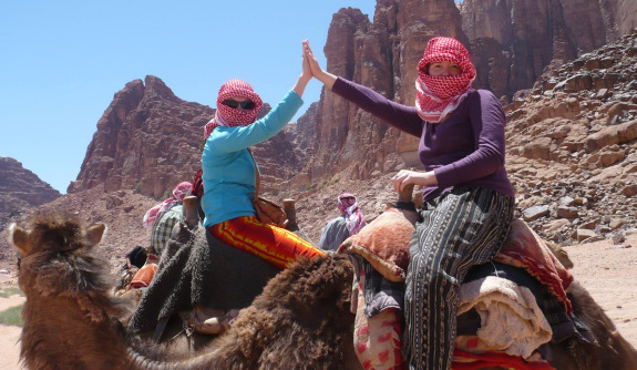 Two students wearing head scarves high-fiving as they ride camels