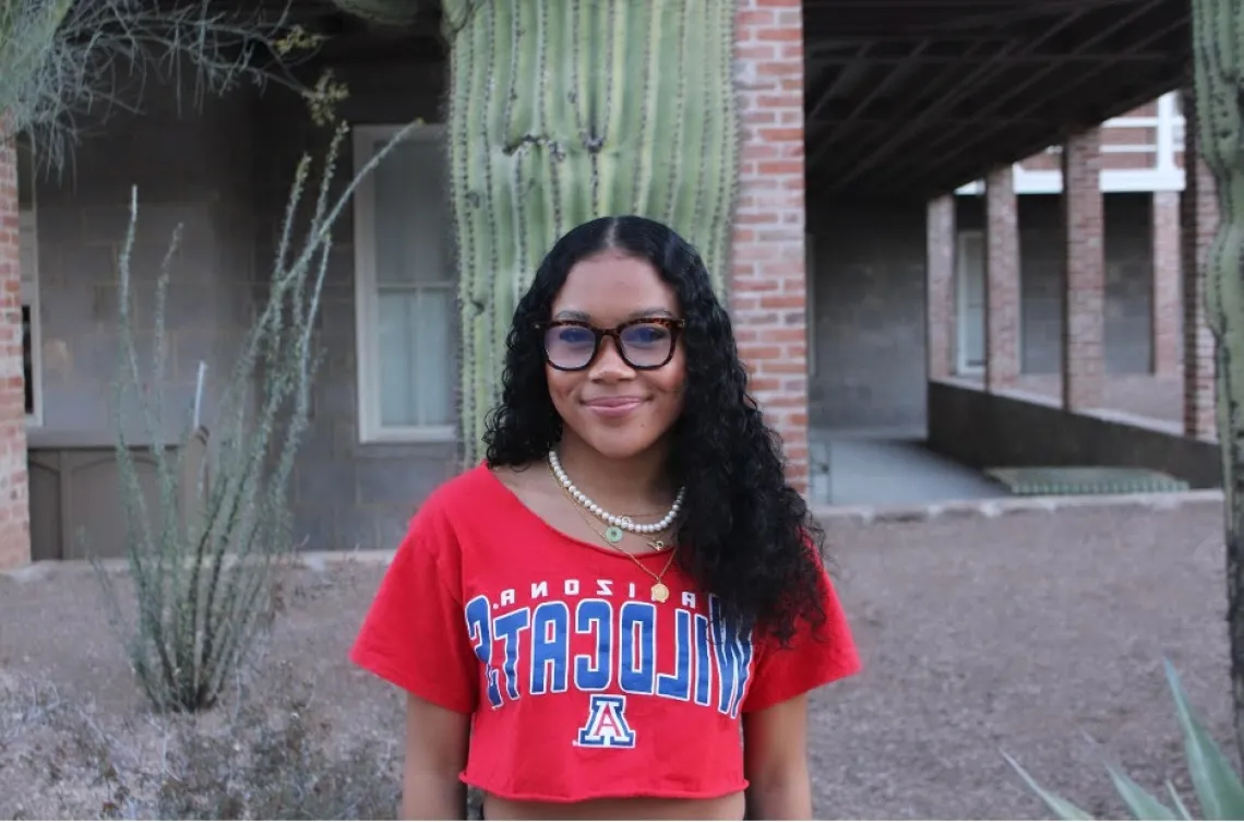 Woman with long dark hair and red shirt standing in front of brick building and a cactus