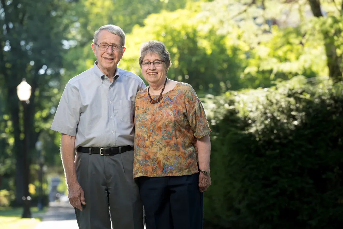 A man and woman stand together outside with sunlit trees in the background