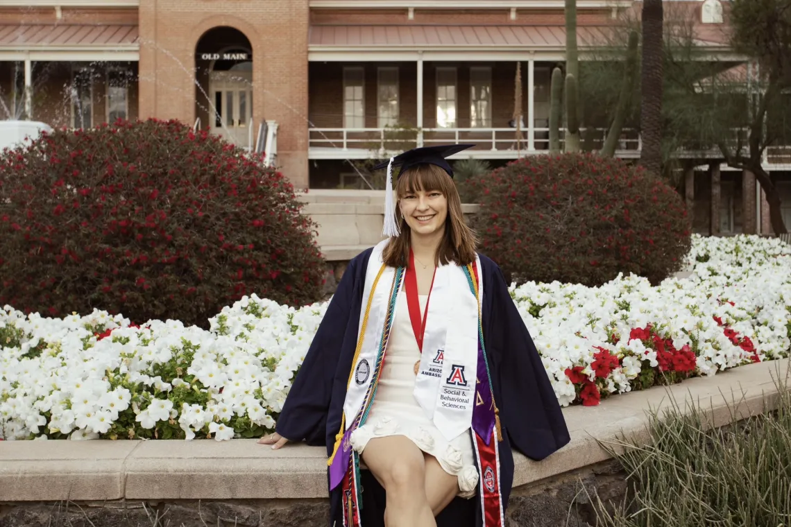 Woman in graduation dress and sandals sits on a brick wall with flowers behind her