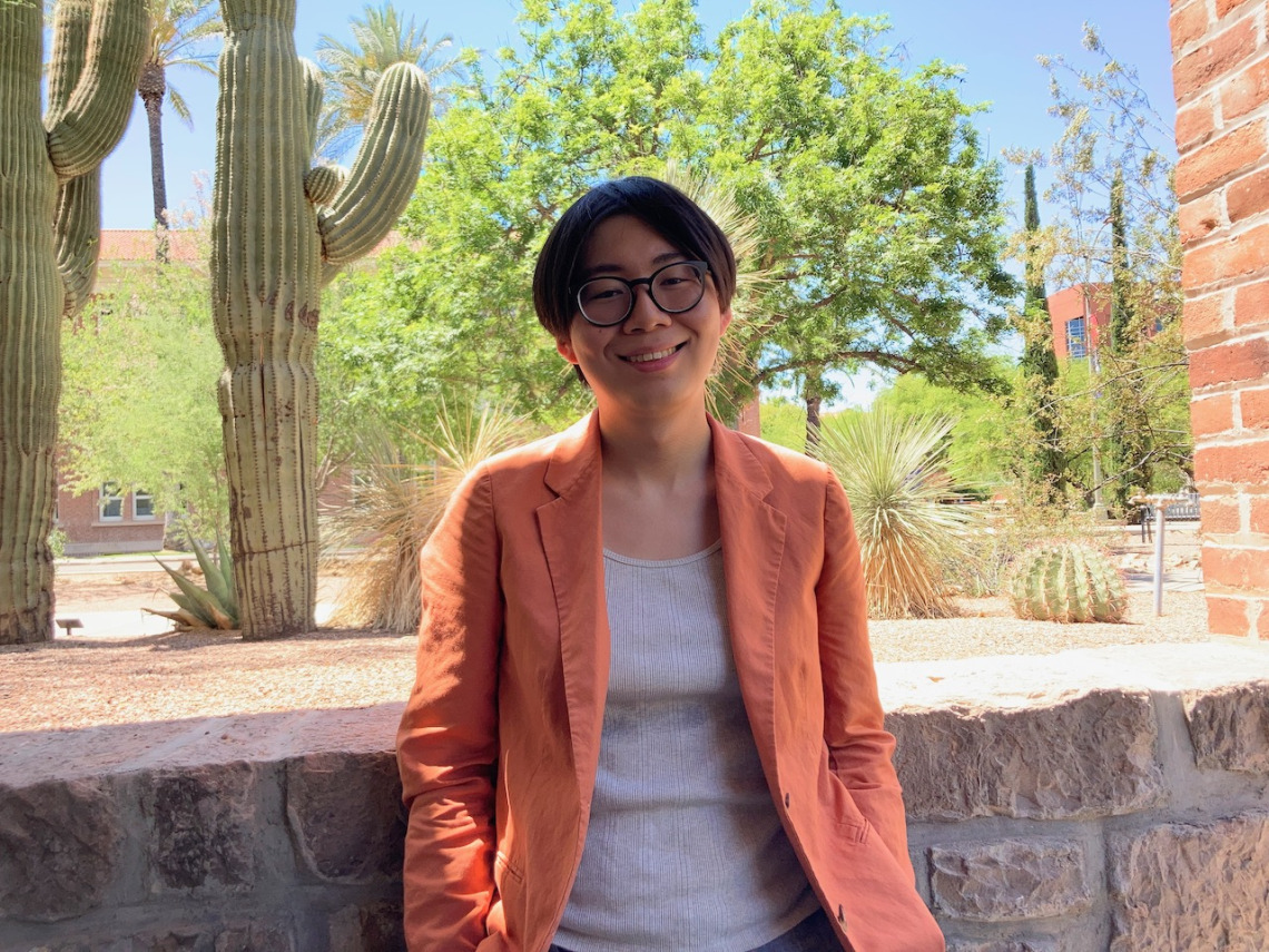 A graduate student with short black hair, glasses and an orange jacket stands outside with a backdrop of cacti and a brick building
