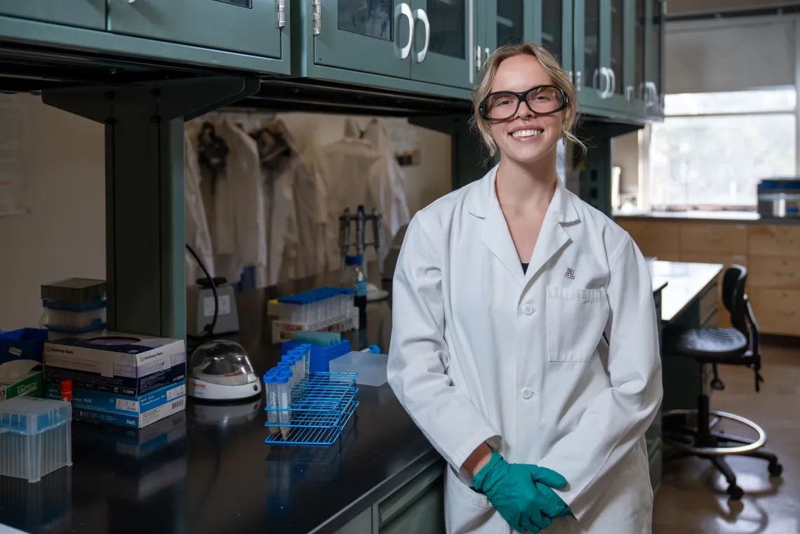 A student with blonde hair, wearing lab glasses and a white lab coat, stands smiling in a lab