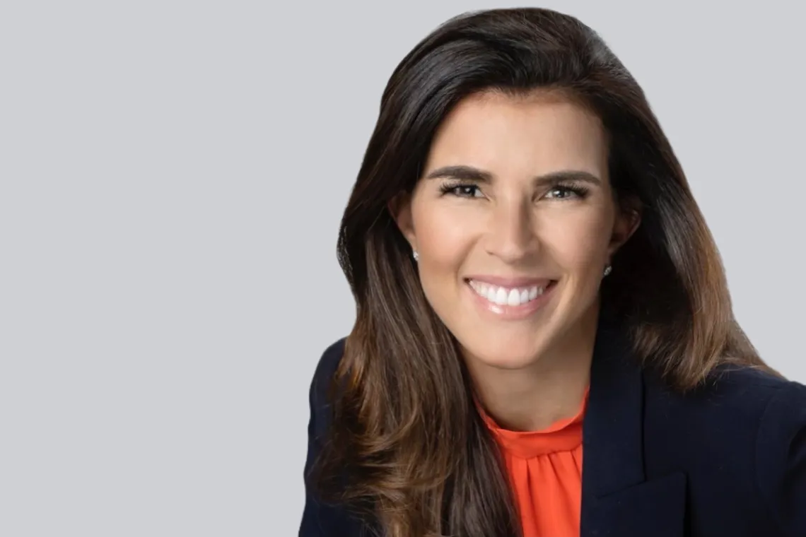 Woman with long brown hair, black jacket and orange blouse smiles in front of a gray background