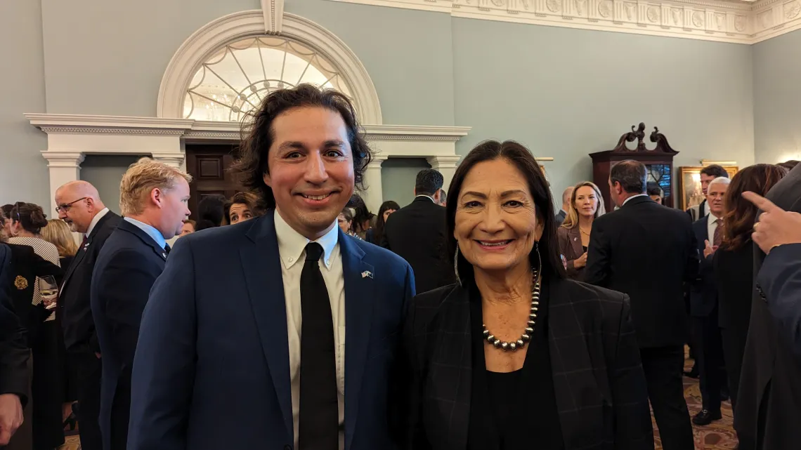 A man in a black suit stands with a woman in a black dress in a large room full of people