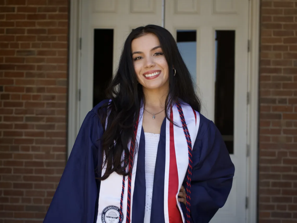 Student with long brown hair, wearing graduation regalia, stands outside in front of a brick building and white doors