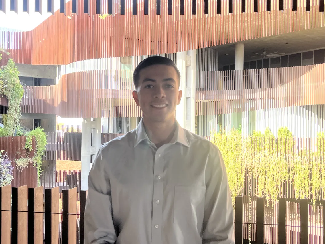 Man in a tan shirt stands outside with greenery behind him