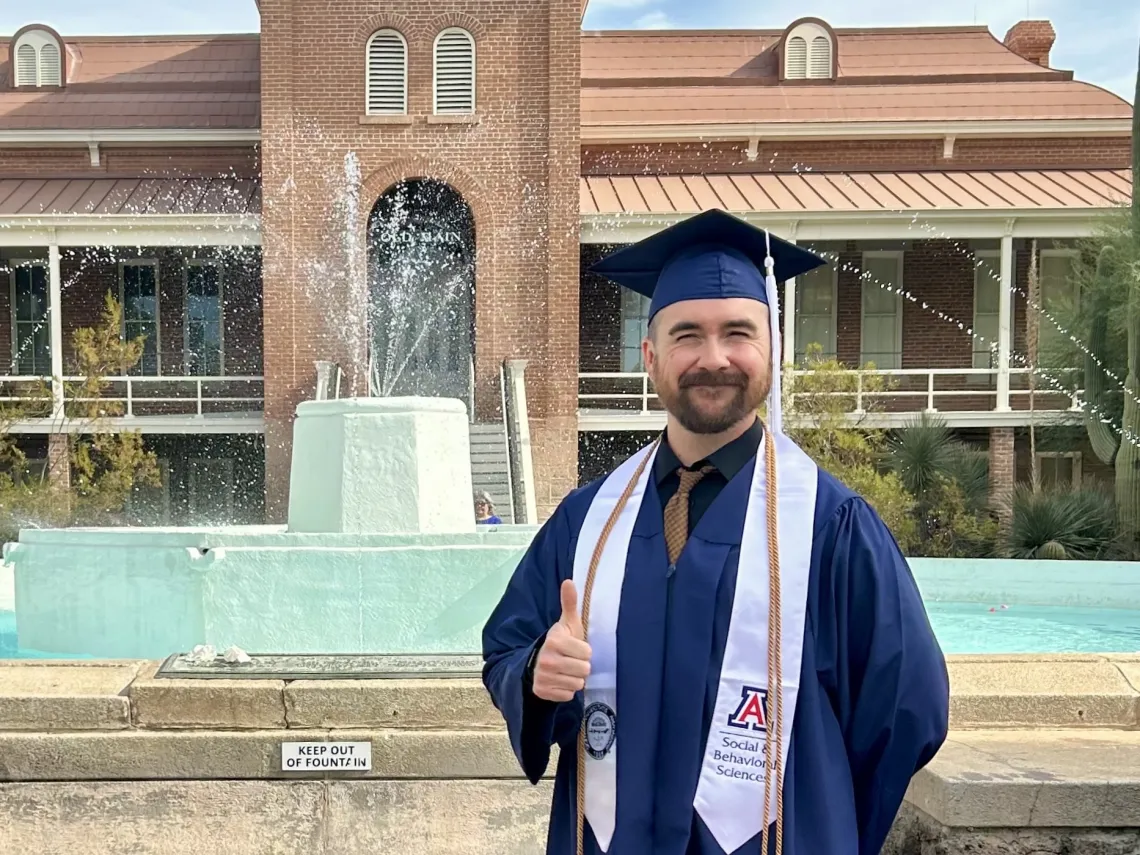 Man wearing graduation cap and gown and giving a thumbs up, stands in front of brick building and fountain. 