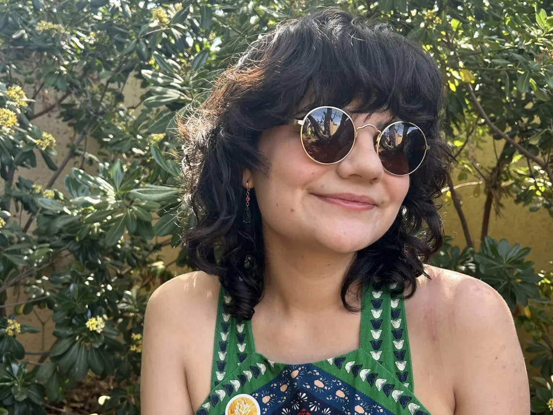 Woman with medium length curly brown hair and wearing sunglasses sits outside