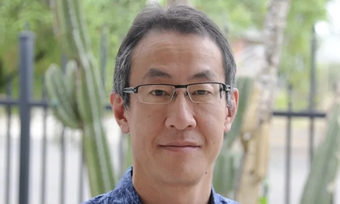 Man with dark hair, glasses and blue shirt stands outside in a wooded area