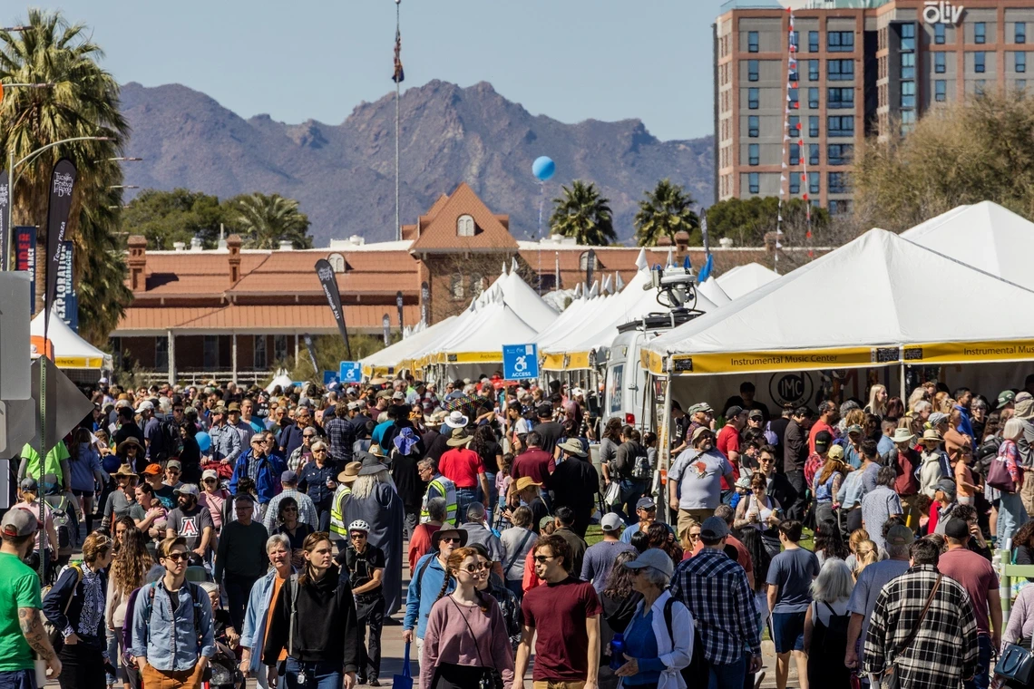 Crowd of people mill around white tents with mountains and palm trees in the background