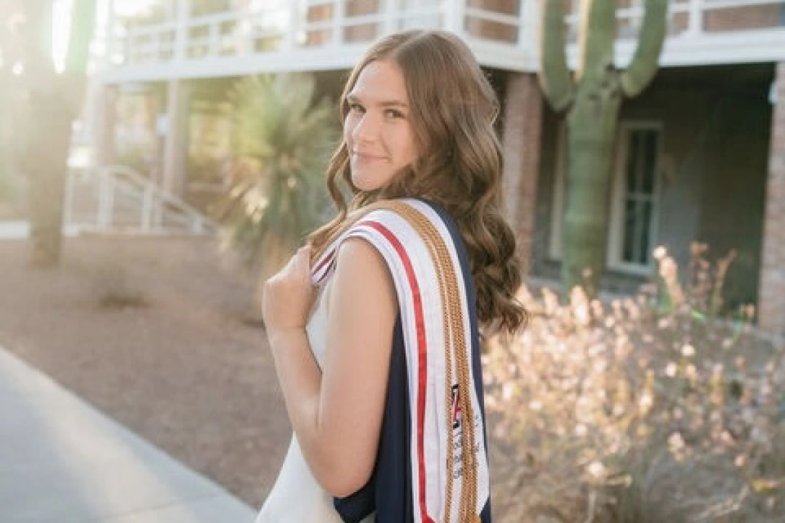 Student with long light brown hair and wearing graduation regalia stands outside in front of a cactus
