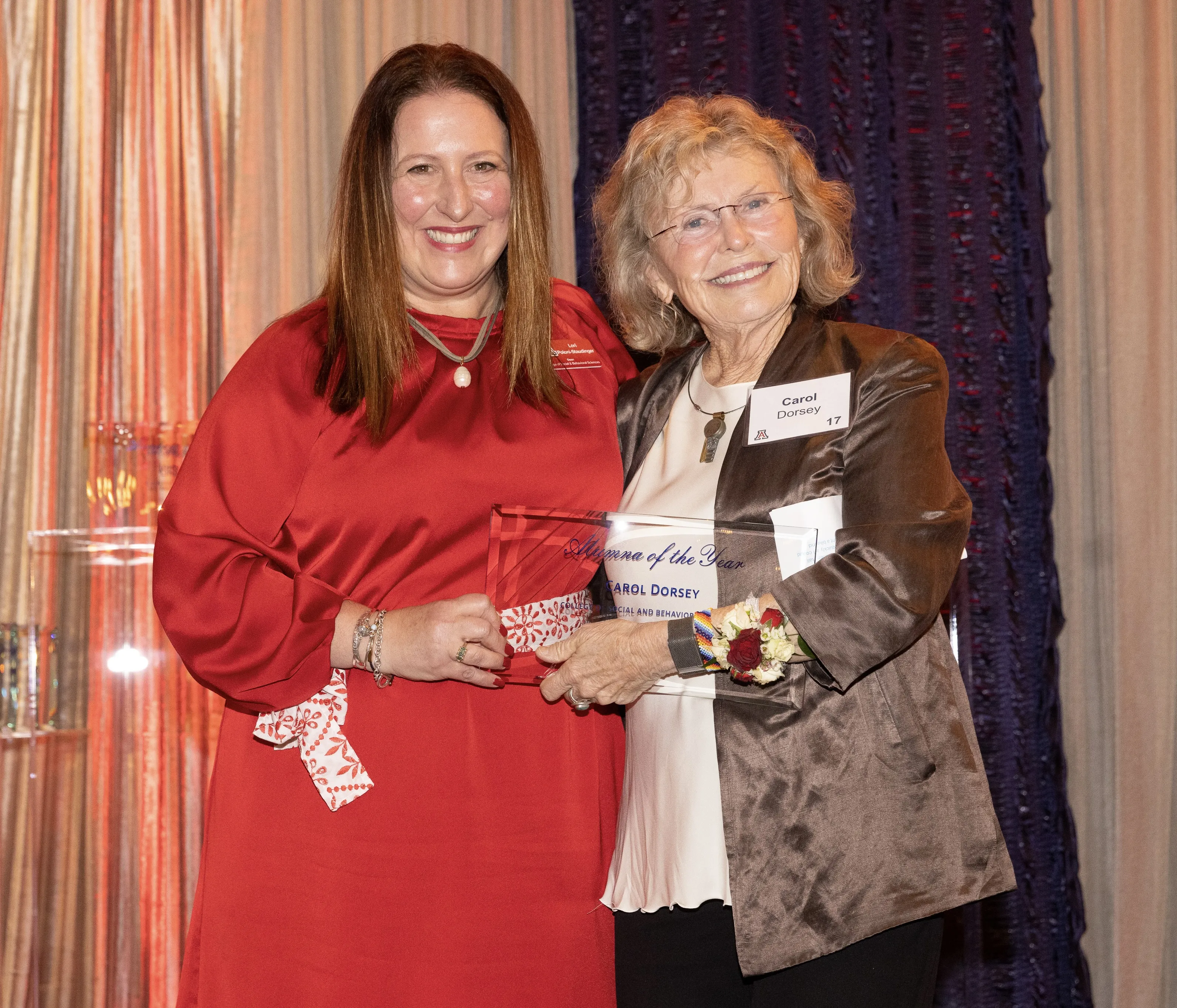 Two women, one in a red dress and one in a brown jacket, stand together, smiling