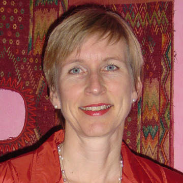 Woman with short blonde hair and red jacket, with a tapestry in the background