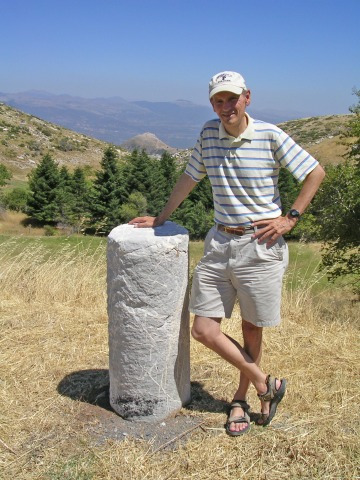 A man in shorts, wearing a baseball cap, stands next to an ancient excavated pillar