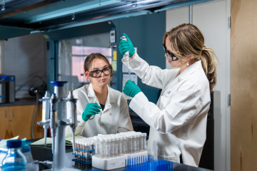 Two women stand in a lab, wearing white coats and working on a project