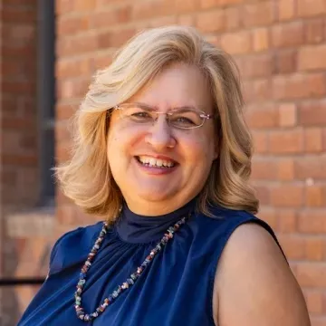 Woman with shoulder-length blonde hair and blue top sits outside against a brick wall