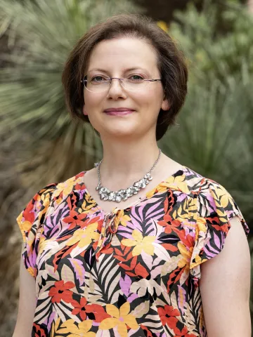 Woman with short brown hair, glasses and floral shirt stands outside smiling