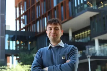 Man in blue sweater stands outside in front of a building, with his arms crossed and smiling