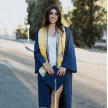 Woman with dark hair, standing in a street wearing a blue graduation gown