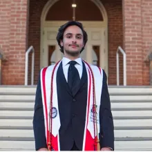 Man with dark hair stands outside in front of an old building, wearing a blue graduation gown