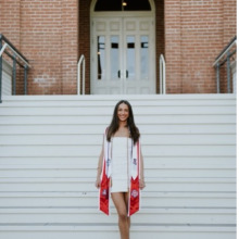 Woman with long dark hair standing on white steps of old building in a graduation gown