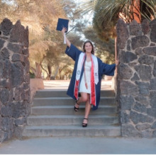 Woman standing on stone steps, holding a diploma and wearing a blue graduation gown