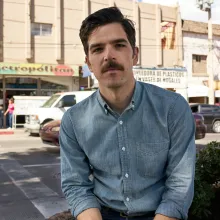 Man with denim shirt and mustache sits outside on a busy street