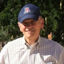 A man wearing a plaid shirt and University of Arizona ball cap stands outside