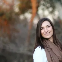 Woman with long brown hair and brown scarf stands outside with trees behind her