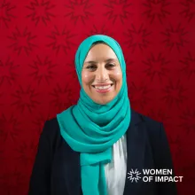 A woman with a turquoise hijab and black jacket sits in front of a red background