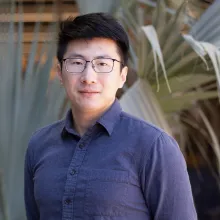 Man with black hair, glasses and a blue shirt stands outside in front of a palm tree