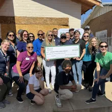 A large group of people wearing sunglasses, stand outside in front of a house under construction. Two of the people hold a large cardboard check showing the gift from Sprouts Foundation