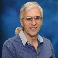 Man with gray hair and glasses smiles in front of a blue background